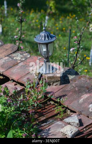 Decorative garden lamp stands on weathered roof tiles surrounded by spring flowers and green meadow, creating a rustic outdoor scene in bright sunligh Stock Photo