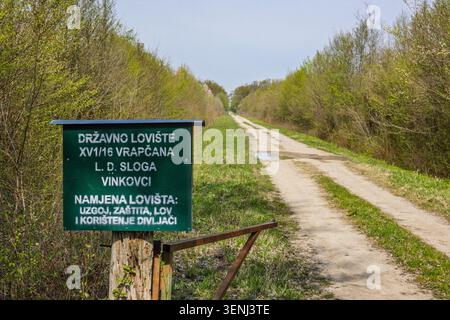 Vinkovci, Croatia - April 10.2026:Green metal hunting sign marks the entrance to a state hunting ground beside a remote dirt road winding through fres Stock Photo