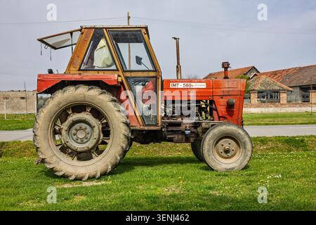 Vinkovci, Croatia - April 10.2026:A vintage red tractor parked on a grassy field beside a rural farmhouse, displaying worn tires, exposed engine and m Stock Photo