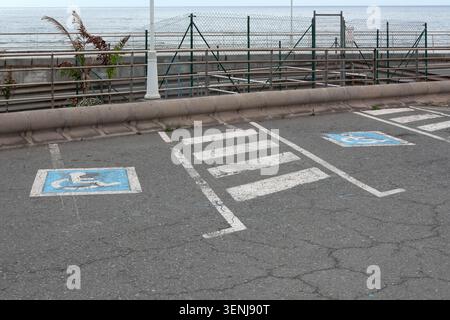 Two blue wheelchair symbols painted inside designated handicap parking spaces separated by white stripes near a coastal promenade and fence line. Stock Photo