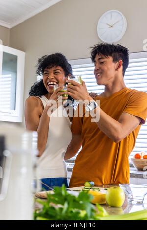 Diverse couple clinking clear glasses and laughing at counter after blender finishing green juice Stock Photo