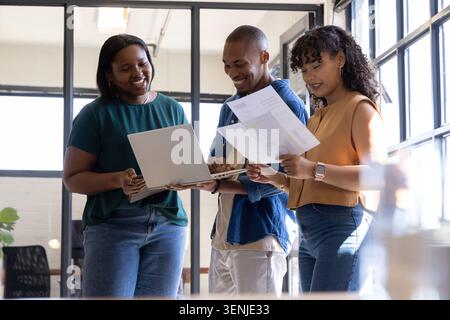 Diverse coworkers reviewing documents and holding open laptop in glass-walled office in workwear Stock Photo