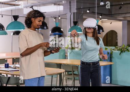 Indian man holding tablet beside female coworker wearing white VR headset interacting in office Stock Photo