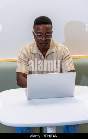 Adult African man sitting in booth at round table wearing clear eyeglasses using silver laptop Stock Photo