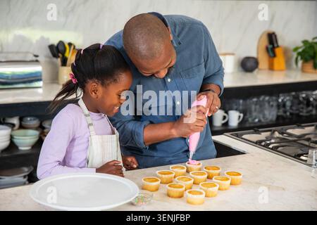 African American father  and  daughter in apron icing cupcakes at counter with pink piping bag Stock Photo