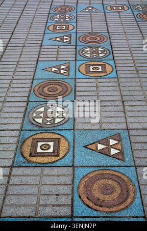 A geometric mosaic pattern integrated into stone pavement tiles features blue, yellow, and brown shapes including circles, triangles, and squares on a Stock Photo