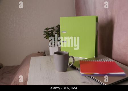 A green folder sits on a wooden desk next to a colorful notebook and a gray mug. A small plant is nearby. The light is soft and warm in the afternoon. Stock Photo