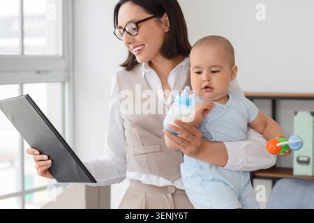 Working mother holding baby and bottle while reviewing documents in a bright office setting, challenges and rewards of modern parenting and career man Stock Photo