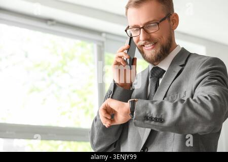 Smiling businessman in gray suit talking on smartphone and checking smartwatch for time management Stock Photo