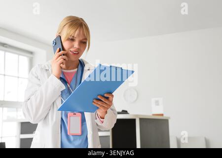 Female doctor or nurse communicating on smartphone while reviewing patient medical records on clipboard in clinic, healthcare professional communicati Stock Photo