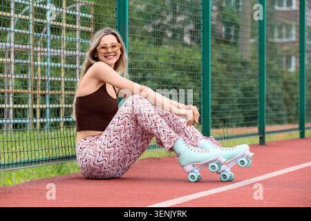 Smiling young woman in retro roller skates and wavy pattern pants sitting by green fence on red track, she has blonde hair and is wearing stylish sung Stock Photo
