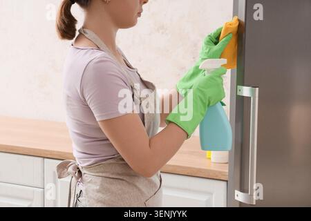 Woman in apron and green gloves cleaning refrigerator with spray bottle and sponge in modern kitchen, she uses a spray bottle and a yellow sponge to w Stock Photo