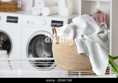 Freshly washed white towels in woven laundry basket on drying rack with washing machines Stock Photo