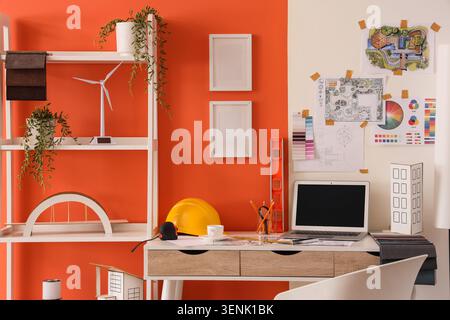 Architectural design workspace with laptop, wind turbine model, and building plans on orange wall Stock Photo