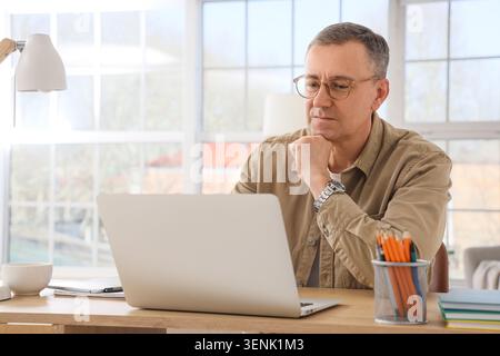 Focused mature man working remotely on laptop at home office desk, thoughtful business professional concentrating on online task Stock Photo