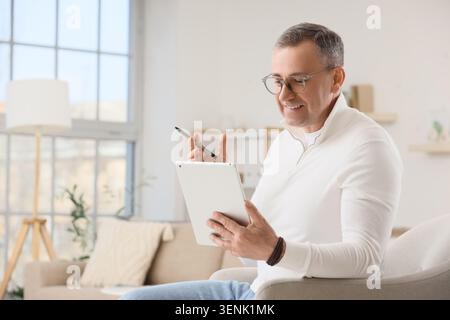 Smiling mature man using digital tablet and stylus pen for online learning or business communication at home, he is casually dressed in a white shirt Stock Photo