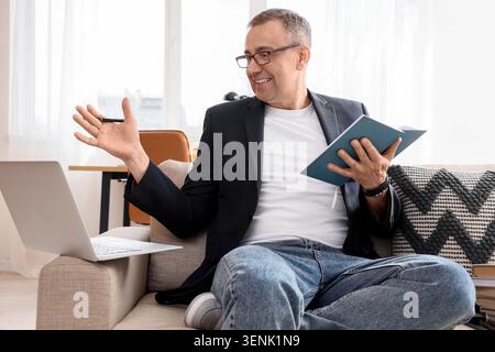 Smiling man in glasses holding notebook and pen, discussing business online with laptop on sofa Stock Photo