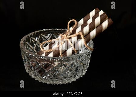 Still life with wafer rolls filled with cream in a crystal vase against a black background. Close-up from the front Stock Photo