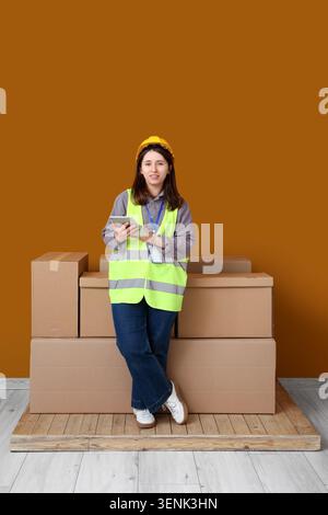 Female warehouse worker in safety vest and hard hat holding tablet next to cardboard boxes Stock Photo