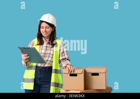 Female warehouse worker in safety vest and hard hat checking inventory on clipboard with cardboard boxes, she stands next to a stack of cardboard boxe Stock Photo