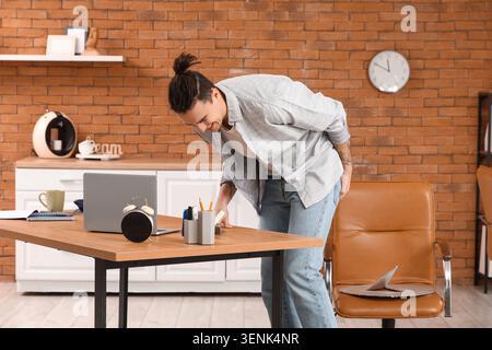 Young man experiencing back pain at home office desk, discomfort from prolonged sitting and poor posture Stock Photo