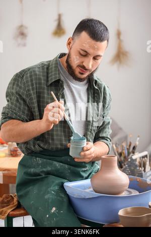 Young male potter mixing light blue glaze for pottery in a creative workshop setting, he is wearing a plaid shirt and a green apron, showing concentra Stock Photo
