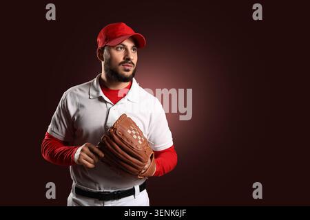 Young adult baseball player in red cap and white uniform holding brown leather glove, looking away thoughtfully, the athlete exhibits focus and concen Stock Photo