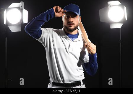 Serious baseball player in uniform holding bat with blue cap and white polo shirt under studio lights Stock Photo