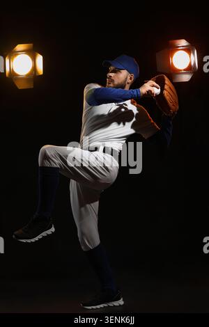 Professional baseball pitcher in uniform with glove and ball on dark background with studio lights Stock Photo