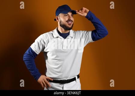 Smiling young man in blue baseball cap and white polo shirt posing confidently on brown background, the man has a beard and is looking to the side wit Stock Photo