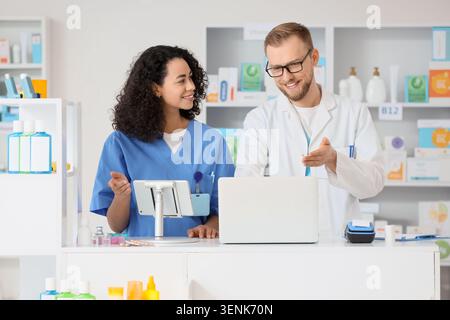 Pharmacist and pharmacy technician collaborating on laptop and tablet for prescription management and customer service, the professionals are wearing Stock Photo