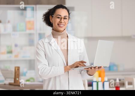 Smiling female pharmacist in white coat using laptop for online pharmacy management and customer service, she stands confidently in a modern drugstore Stock Photo