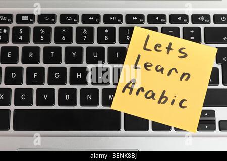 Online arabic language learning with yellow sticky note on laptop keyboard for digital education, the note displays "lets learn arabic" in black handw Stock Photo