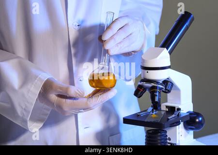 Scientist in lab coat and gloves holding flask with yellow liquid next to microscope for research and analysis, a microscope stands nearby, ready for Stock Photo