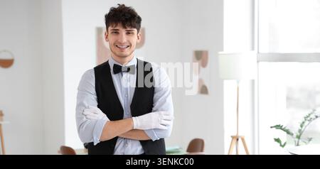 Smiling young waiter in formal vest and bow tie with white gloves posing confidently indoors, this professional male employee is ready for work in the Stock Photo