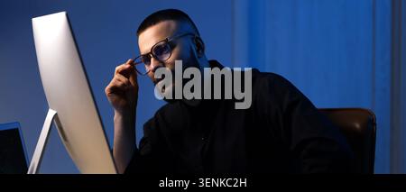 Focused young man adjusting glasses while working on computer in dark office, late night productivity and digital technology Stock Photo