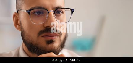 Focused young man in modern eyeglasses with beard, concentrating on work or study, close-up portrait Stock Photo