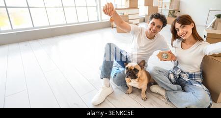 Happy young couple with french bulldog holding new house keys and piggy bank in empty room with moving boxes, the man holds up house keys with a house Stock Photo