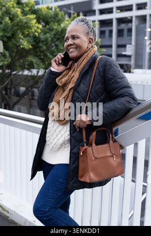 Senior African American woman leaning on railing on bridge, talking on phone, wearing quilted coat Stock Photo