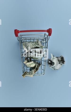 Metal cart holds crumpled bills. One bill lies fallen beside it. Minimalist blue backdrop highlights the scene. No people, no animals, no plants. Idea Stock Photo