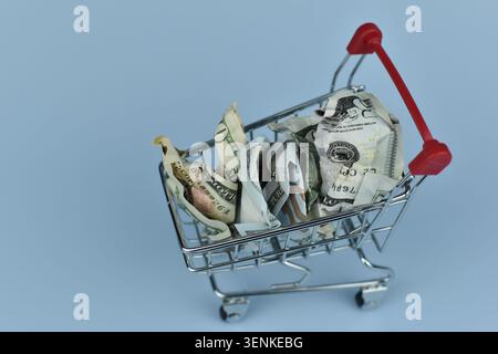 Metal cart holds crumpled bills. One bill lies fallen beside it. Minimalist blue backdrop highlights the scene. No people, no animals, no plants. Idea Stock Photo