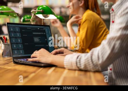 Student using AI study assistant interface typing on laptop at library table, near classmates and green lamps. Modern automation tools in education and online learning. Stock Photo