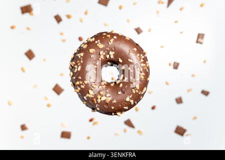 A single, tempting chocolate-covered donut sprinkled with chopped peanuts and surrounded by scattered chocolate pieces against a white background Stock Photo