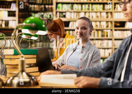 Portrait of smiling student looking at camera from library study table, sitting near classmates for academic study environment. Young woman showing positive learning in reading room. Stock Photo
