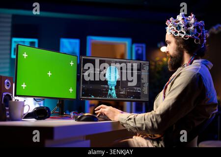 Man with EEG headset on writing code on green screen PC to transfer mind into virtual world, becoming one with AI. Stock Photo
