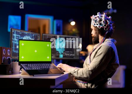 Focus on green screen laptop with man in blurry background using EEG headset to transfer mind into virtual world, close up shot of mockup device next to scientist using tech to gain superintelligence. Stock Photo
