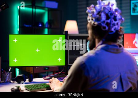 Man with EEG headset programs brain transfer into green screen computer virtual world, merging with artificial intelligence, IT specialist at home using neuroscience and mockup PC to gain digital sou. Stock Photo