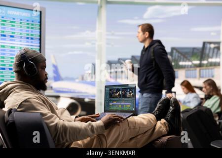 Black videographer reviewing film montage and editing in post production, seated in airport lounge before boarding. International travel with nomadic lifestyle, work anywhere concept. Stock Photo