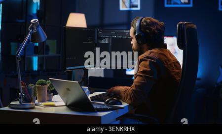 Engineer typing code in apartment office, using javascript programming language to develop software application. IT admin using multimonitor desktop setup and notebook to finish tasks, camera A. Stock Photo