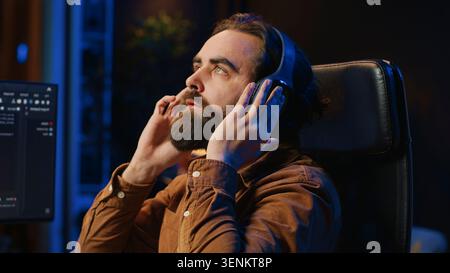 Cheerful programmer listening music while doing web development during remote job shift in neon lit home office, finishing coding project. IT expert enjoying audio podcast, working on PC, camera A. Stock Photo
